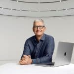 Tim Cook, CEO da Apple, sentado numa mesa moderna com um portátil MacBook, refletindo sobre inovação tecnológica e políticas de imigração nos EUA.