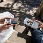 Duas pessoas a tirar selfies com smartphones ao ar livre, sentadas em escadaria com sombra de árvores, destacando a tecnologia móvel moderna.