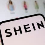 Imagem de um smartphone exibindo o logo da Shein, com fundo de telas de aplicativos de moda infantil.