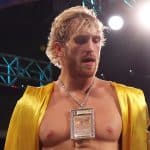 Imagem de um homem com cabelo loiro, sem camisa, usando uma jaqueta amarela e um colar com uma carta Pokémon pendurada.